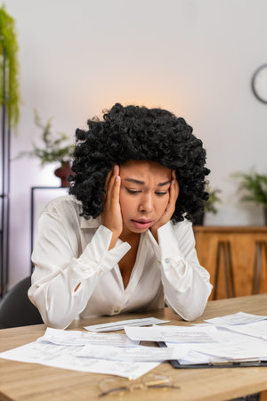 African American businesswoman at home office studies overdue bills fearing debt and high taxes. Freelancer girl at table counts receipts invoices seeking cuts to protect savings and avoid bankruptcyの写真素材
