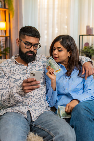 Indian couple man and woman sitting on home sofa holding money dollar cash and smartphone while planning purchases and family budget. Discussing future expenses, savings and income from salary indoorsの写真素材