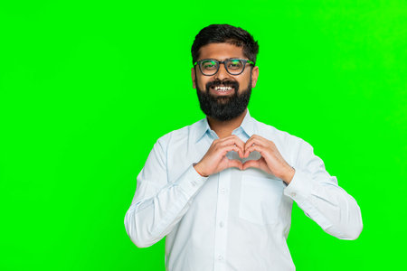 Man forms heart shape with hands near chest smiles gently, expressing love care warmth in friendly gesture. Guy isolated on green chroma key background showing kind emotional messageの写真素材