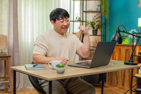 Asian man sitting at home table works on laptop, finishing project, clenching fist, smiling with satisfaction. Chinese guy proudly celebrates project success, energized and motivated by achievement.の写真素材