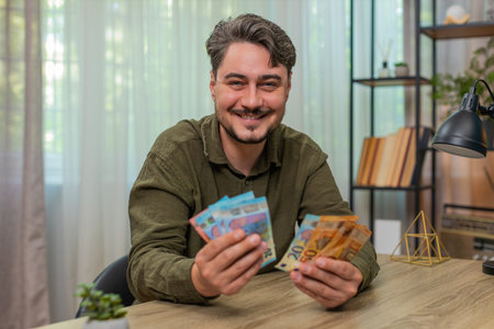 Middle-aged man at home counts cash smiling pleased by financial success feeling secure and proud. Young adult guy at table flips euro bills stacking money appreciating wealth gains after hard workの写真素材