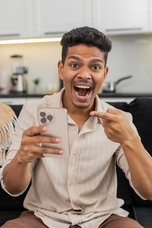 Man at home watches video on smartphone feels shock laughs smiles excited by news. Guy on sofa shares link in messenger sends to friends writes short reply and posts reaction emojiの写真素材