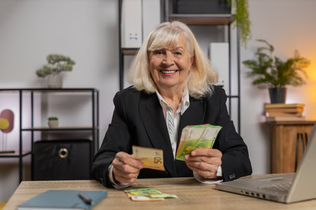 Excited senior businesswoman counting cash money checking salary enjoy richness at workplace. Happy...