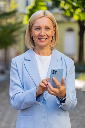Portrait of happy mature businesswoman standing on the city street. Smiling Caucasian woman employer in formal suit using the smartphone and looking at the camera. Headshot of positive female manager.の写真素材