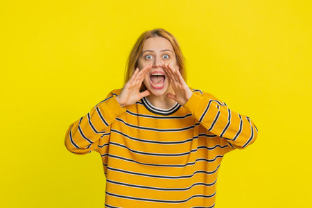 Expressive Caucasian young blonde woman holding hands near mouth loudly screaming, shouting,...