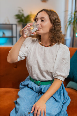 Satisfied young Caucasian woman sitting at home on sofa couch leading healthy lifestyle, drinking a glass of pure water, smiling. Drink daily norm of water. Girl smiling at camera at home. Verticalの写真素材