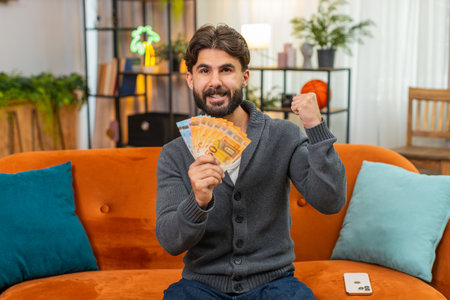 Man at home counts cash banknotes plans travel budget thinks where to spend salary, earnings, savings. Guy holds money stack dreams of trip spend plan feels very happy confidentの写真素材