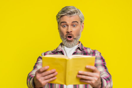 Mature man opens book with eager hope, showing bright surprise and stunned excitement. Middle-aged guy on yellow background shows amazement shocked wow reaction, suggesting hidden intrigue in pagesの写真素材