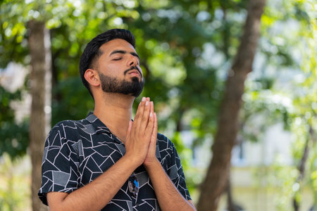Bearded Indian man praying with closed eyes. Young guy clasping hands wishing luck in urban sunny...