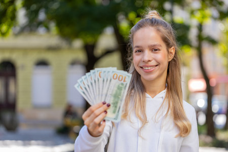 Portrait of successful rich Caucasian girl holding fan of dollars, shows finances with thumbs up gesture on city street.の写真素材