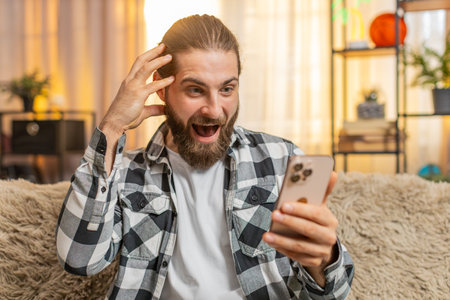 Young man sits at home rapidly inputting text on phone, seeking perfect discount. Guy sitting on sofa exclaiming wow expression while discovering a highly beneficial offerの写真素材