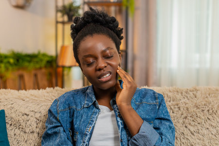 African American young woman painfully touches her cheek and closes her eyes from a sharp toothache,...