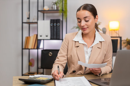 Young businesswoman at home office checks receipts and invoices enters numbers on calculator and totals month. Freelancer girl at table nods satisfaction writes summary and saves report to folderの写真素材