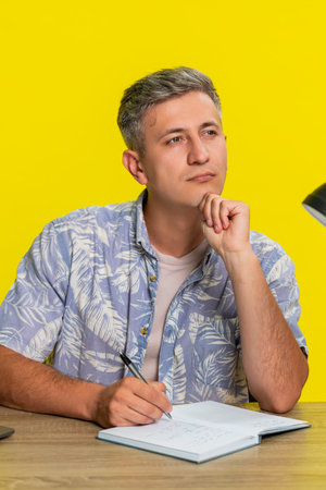 Middle-aged man sitting at table with laptop and notebook, writing notes with serious thoughtful...