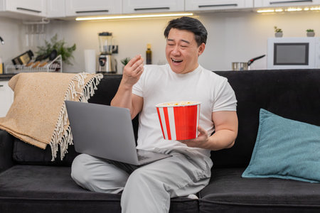 Asian man at home streams comedy on laptop holds bowl of popcorn laughs hard and smiles wide feels...