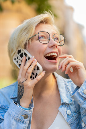 Happy Caucasian young woman in pleasant conversation on smartphone, enjoying talking with friend outdoors. Girl in trendy clothes on urban city street. Town lifestyles.の写真素材