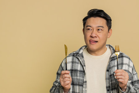 Ready to eat. Hungry Asian middle-aged man waiting for serving dinner dishes with restlessness holding cutlery fork knife, will appreciate delicious restaurant meal on beige backgroundの写真素材