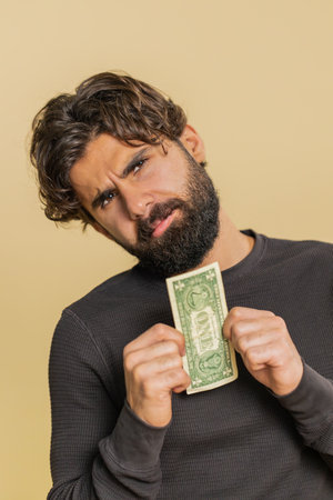 Young man with insufficient amount of money, holding one dollar banknote. Financial crisis. Bankruptcy. Poverty and destitution. Adult guy standing on beige background.の写真素材