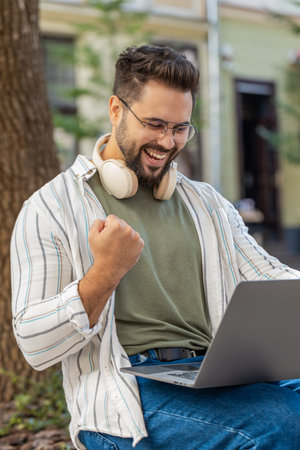 Overjoyed happy young man working on laptop netbook screaming in delight triumph winner gesture celebrate success game win outdoors. Happy adult guy in glasses sitting on street in city park.の写真素材