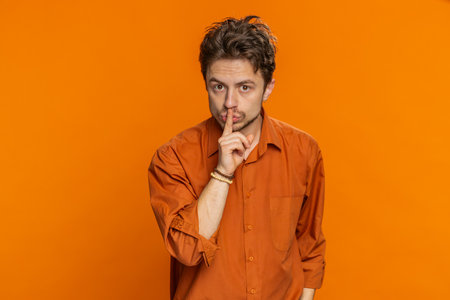 Caucasian man presses index finger to lips makes silence gesture sign do not tells secret, stop talk gossip, confidential privacy. Handsome young guy isolated on orange backgroundの写真素材