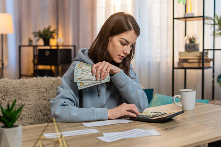 Young Caucasian woman sitting on sofa couch counting money dollar while reviewing bills. Adult girl checks receipts and uses calculator at table checking expenses and budget with focused expression.の写真素材