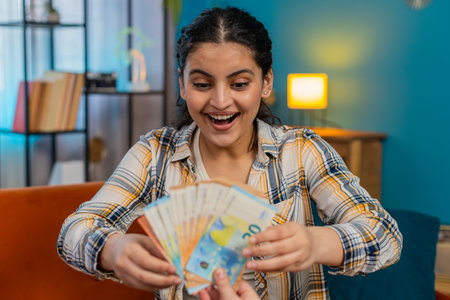 Indian young woman experiencing sudden happiness, joyfully celebrating on sofa receiving plenty of cash. Arabian girl sincerely delighted, smiling widely, enthusiastically showing victory winの写真素材