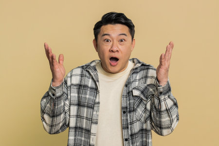 Excited amazed winner Asian man raising hands in surprise looking at camera with big eyes shocked by sudden victory celebrating success achievement good news. Guy on beige backgroundの写真素材