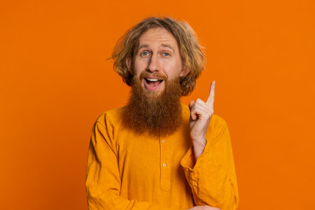 Eureka. Inspired Caucasian man pointing finger up with open mouth, having good idea, plan, startup, showing inspiration motivation gesture, problem solution. Bearded guy isolated on orange backgroundの写真素材