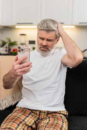 Middle-aged man at home on smartphone sees failed play game or job denial news sighs and shakes head no. Mature guy on sofa lowers gaze breath heavy pauses and tries to calm emotion and accept resultの写真素材