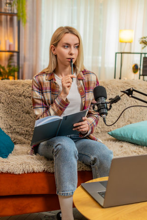Blogger woman prepares to record podcast vlog on laptop while sitting on home sofa, adjusting mic...