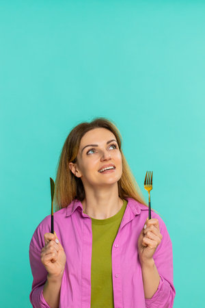 Middle-aged woman holds fork and knife with eager facial expression clearly showing she is hungry...