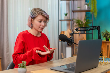 Woman recording video interview at laptop sitting at home table, speaking seriously in studio...