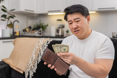 Asian man at home opens wallet sees few small dollar bills frowns sighs and sets new saving goal....