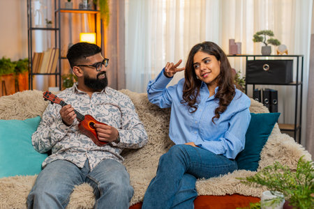 Couple man woman sitting on home sofa, guy playing toy ukulele while girl dances and both laugh with...