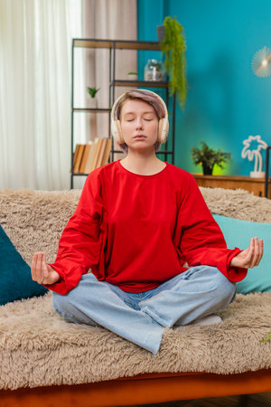 Woman quietly meditating at home using wireless headphones, breathing deeply with peaceful...