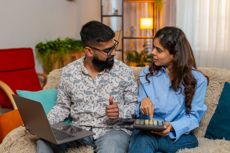 Indian couple man and woman sitting on home sofa with laptop and calculator calmly discussing expenses and future savings. Young family working on home budget plan with focus and understanding indoorsの写真素材