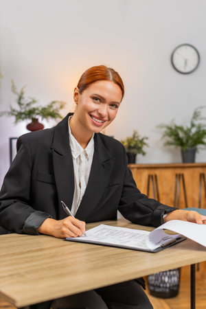 Young businesswoman at home office turns pages of contract reading each line and signing with hand. Freelancer girl at table finalizes paperwork sealing deal satisfied with legal clarity project startの写真素材