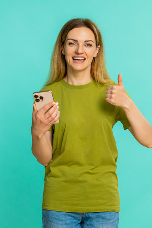 Young woman smiles brightly while looking at smartphone and showing thumbs up to camera expressing...