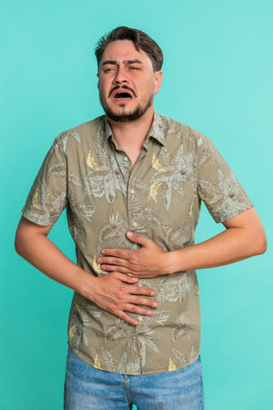 Middle-aged man holds stomach leans forward with tense expression indicating sudden pain or discomfort. Young adult guy isolated on blue background suffering from unexpected abdominal cramp or illnessの写真素材