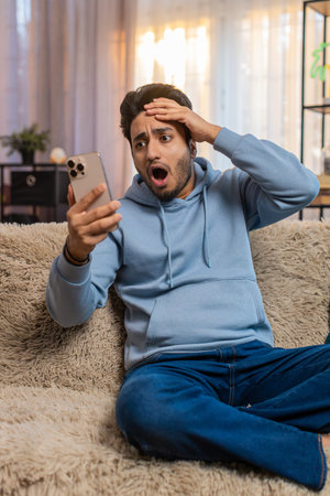 Shocked sad young man using smartphone, reading negative message, feeling annoyed sitting on sofa in...
