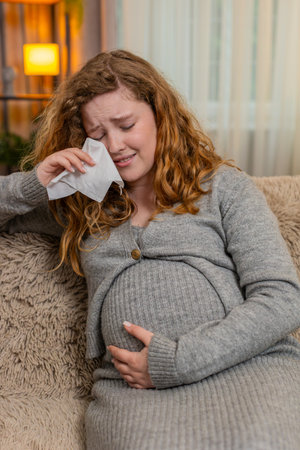 Pregnant woman sits on home sofa gently holding her belly, wiping tears with emotional expression,...