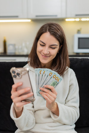 Young woman at home counts dollar cash checks rate in phone app smiles and plans big buy goal....