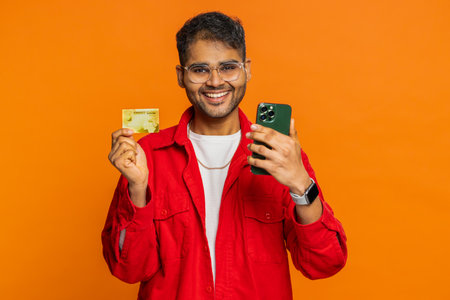 Happy smiling Indian man showing credit card and smartphone while transferring spending money,...