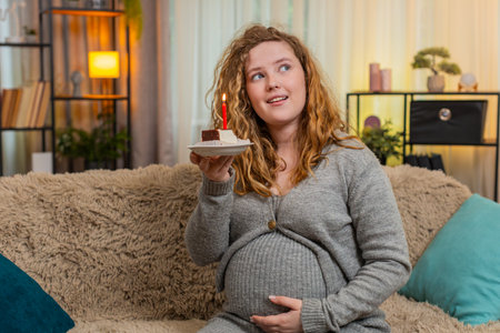 Pregnant woman sits on home sofa, blowing out birthday candle on cake making a wish then. Girl embraces sweet moments, celebrating life and finding happiness in small pleasuresの写真素材