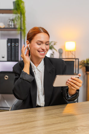 Young businesswoman at home office watches smartphone music clip using wireless headphones dances in rhythm, laughs showing joy relaxing. Freelancer girl at table free moves to beat celebrating momentの写真素材