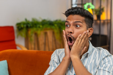 Man at home grabs cheeks and says wow after reading positive news on smartphone and smiles. Guy on sofa feels surprise opens mouth raises eyebrows claps hands and shows joyful reaction.の写真素材