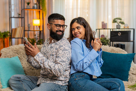 Couple man and woman sitting on home sofa back to back with smartphones, smiling and watching funny...