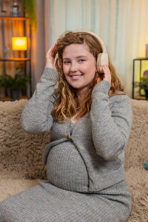 Pregnant young woman listens to music on headphones at home, gently touching her belly, dancing with careful steps and positive mood. Girl enjoys the rhythm, feeling love, connection, and happiness.の写真素材