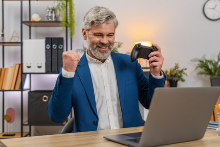 Middle-aged businessman at home office lifts joystick skyward after laptop game victory cheering...