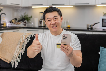 Asian man at home pays with smartphone and credit card waits for confirmation then smiles with joy....
